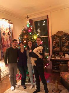 Four friends stand in front of a Christmas tree lit with multicolored lights; the man standing on the far right is holding a white dog.