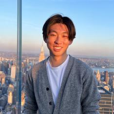 Joshua Yang ’25 stands in late afternoon sun from somewhere high up in New York City, with the cityscape in the background.