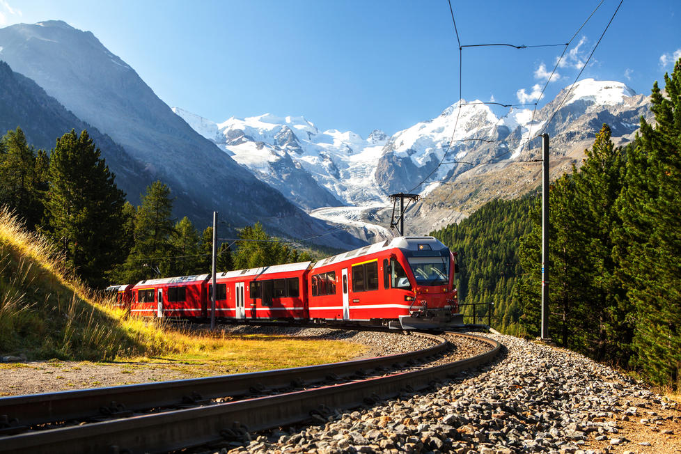 A bright red train rounds the corner of the tracks and the Swiss Alps are visible in the background