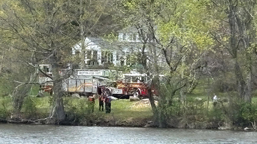 A fire truck is parked on the grass next to a lake, a white house is in the background.