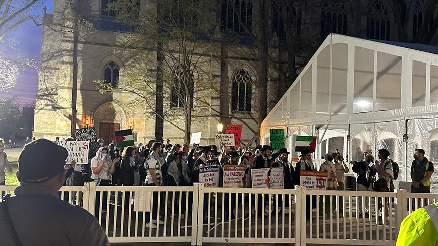 People hold signs behind a white fence with the University Chapel in the background.