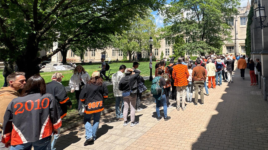 A long line of alumni in orange Reunions gear wait in a line on a stone path.