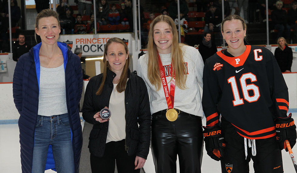 Four women stand on the ice, with one wearing a gold medal.