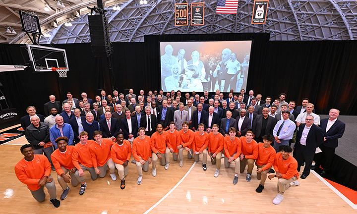 A large group of alumni stand behind the current men's basketball team, who are on one knee. In the background is a large screen showing photos of Pete Carril.