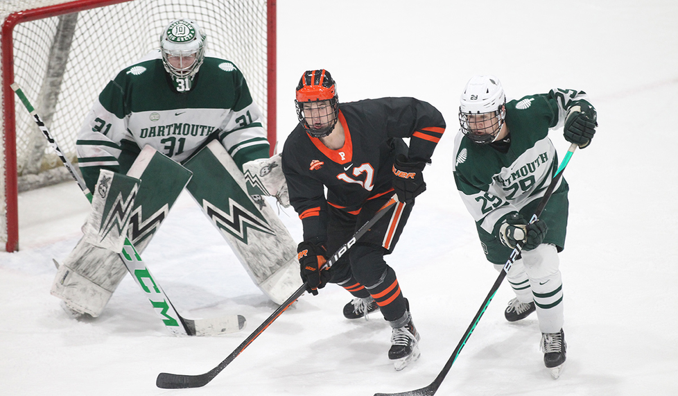 A Princeton men’s ice hockey player skates between a Dartmouth player and the Dartmouth goalie, with the goal in the background. 