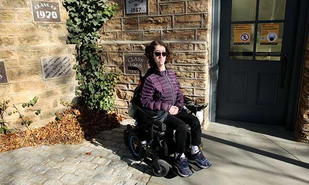 This is a photo of Naomi Hess ’22 in her motorized wheelchair in front of a Princeton campus building. Stone plaques in the wall behind her read "Class of 1970" and "Class of 1968."