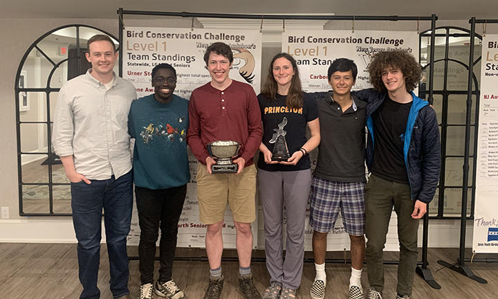 Six students in the winning Princeton team face the camera, with two in the center holding trophies.