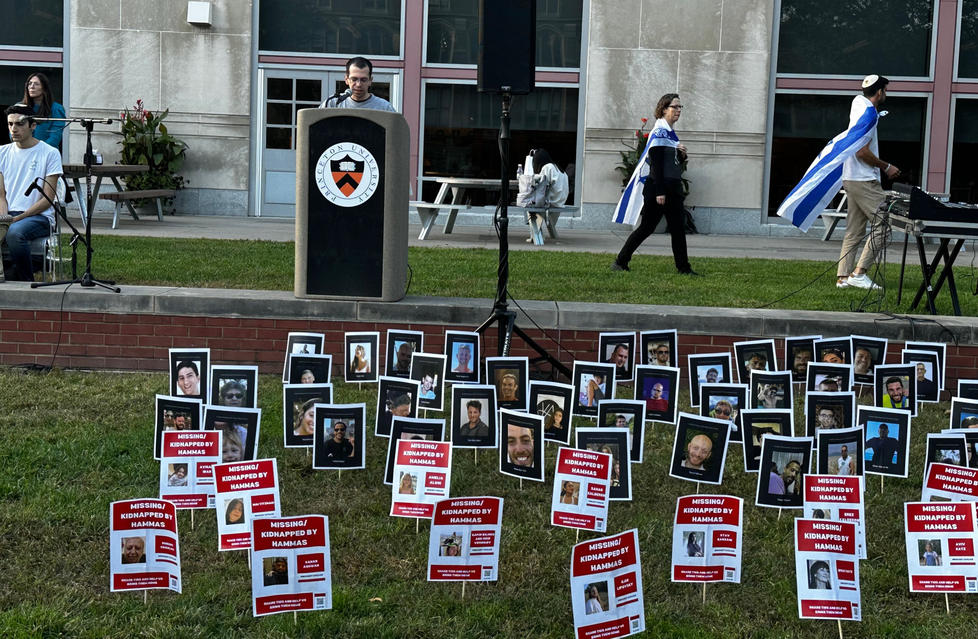 A man speaks at a podium bearing the Princeton seal; in front of him, about 30 photos on stakes are in the lawn. 