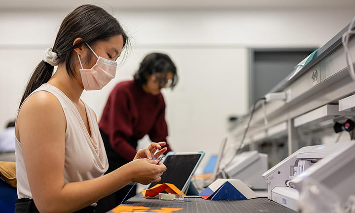 A student wearing a face mask folds orange pieces of paper; another student and a laptop computer are visible in the background.