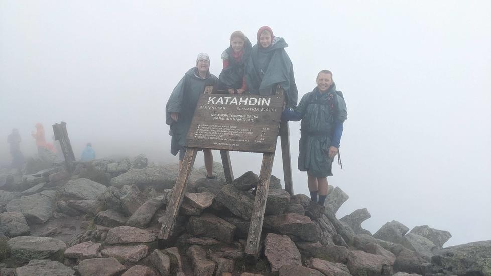 Four people in rain jackets cluster in thick fog around a wooden sign reading “Katahdin,” perched on a pile of rocks.