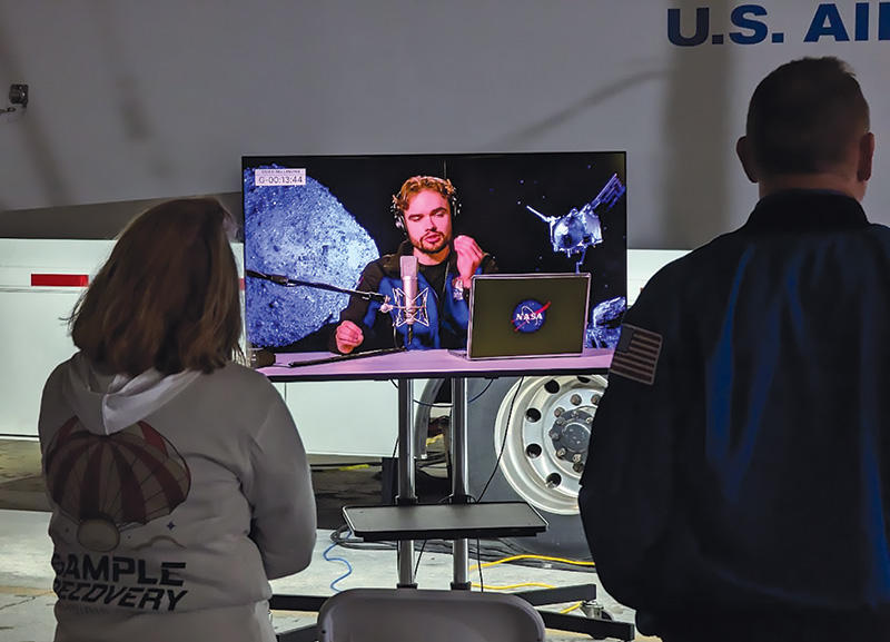 Two people, backs turned, watch Tralie speak on a TV screen.