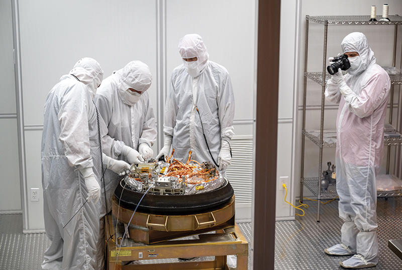 Four people in white hazmat suits stand around the sample from asteroid Bennu as it's unpacked 