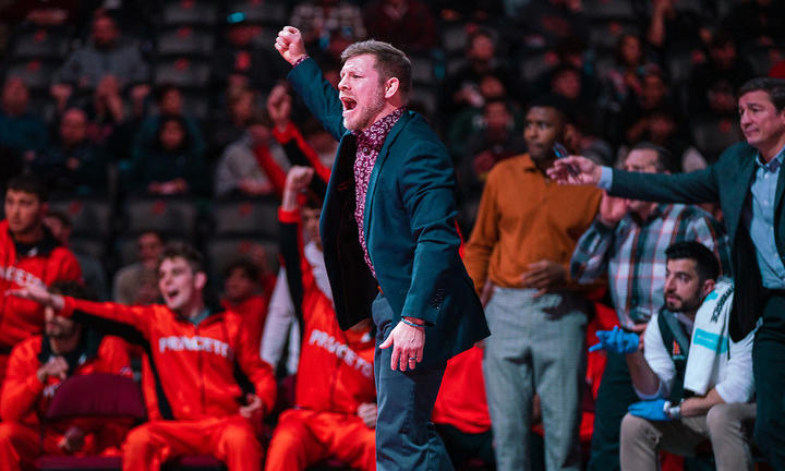 Joe Dubuque yells while raising his fist in the air on the sidelines of a wrestling match; a crowd cheers behind him.